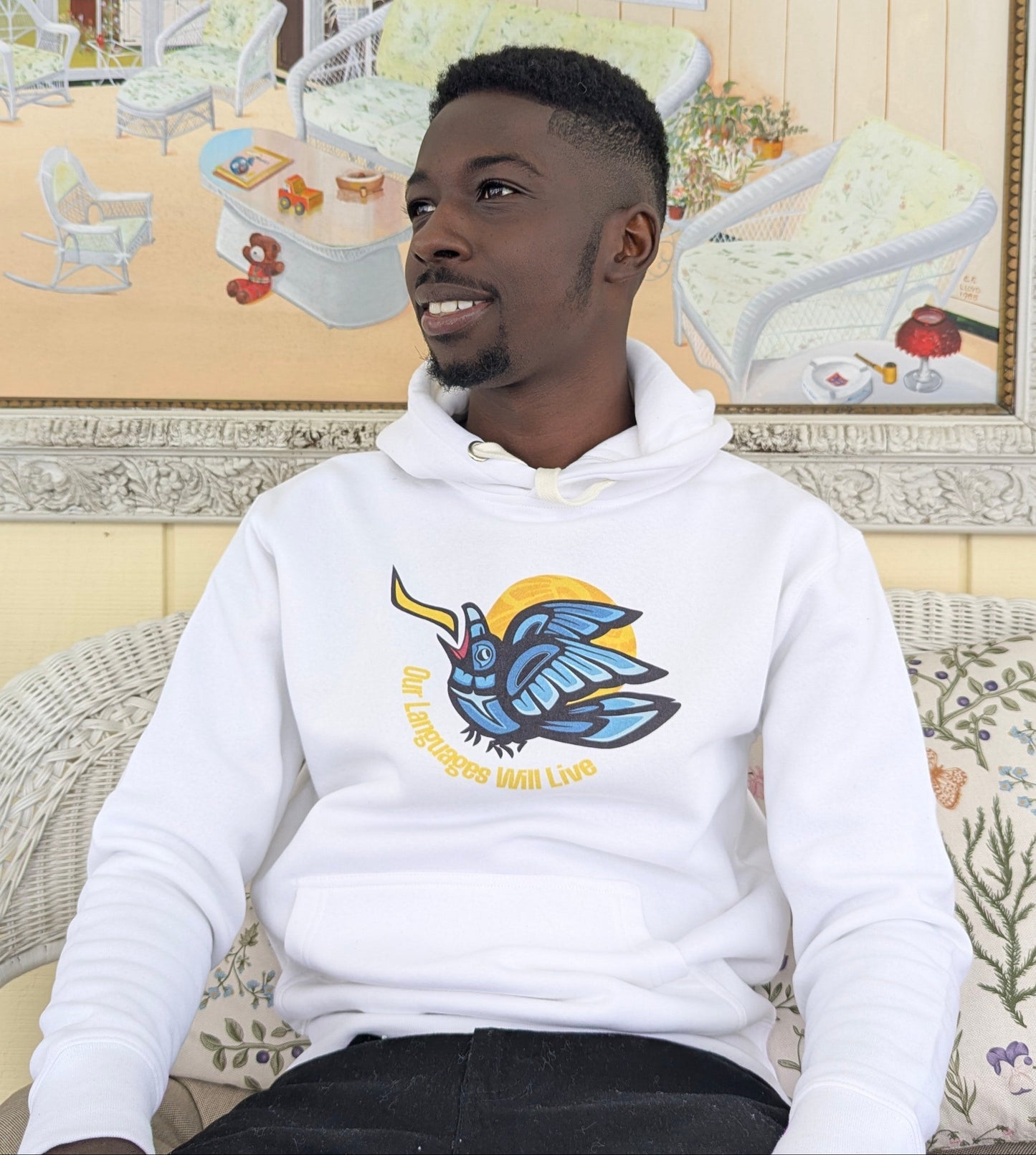 Young Nigerian man wearing a white hoodie with a blue and yellow formline raven illustration, in a room with framed pictures on the wall.